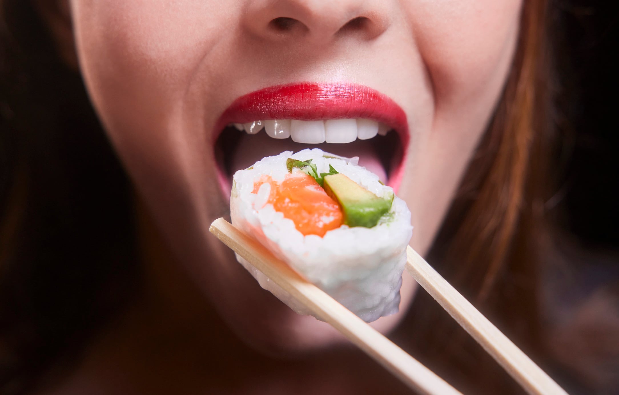 Close-up of woman eating salmon and avocado rice sushi piece on chopsticks.