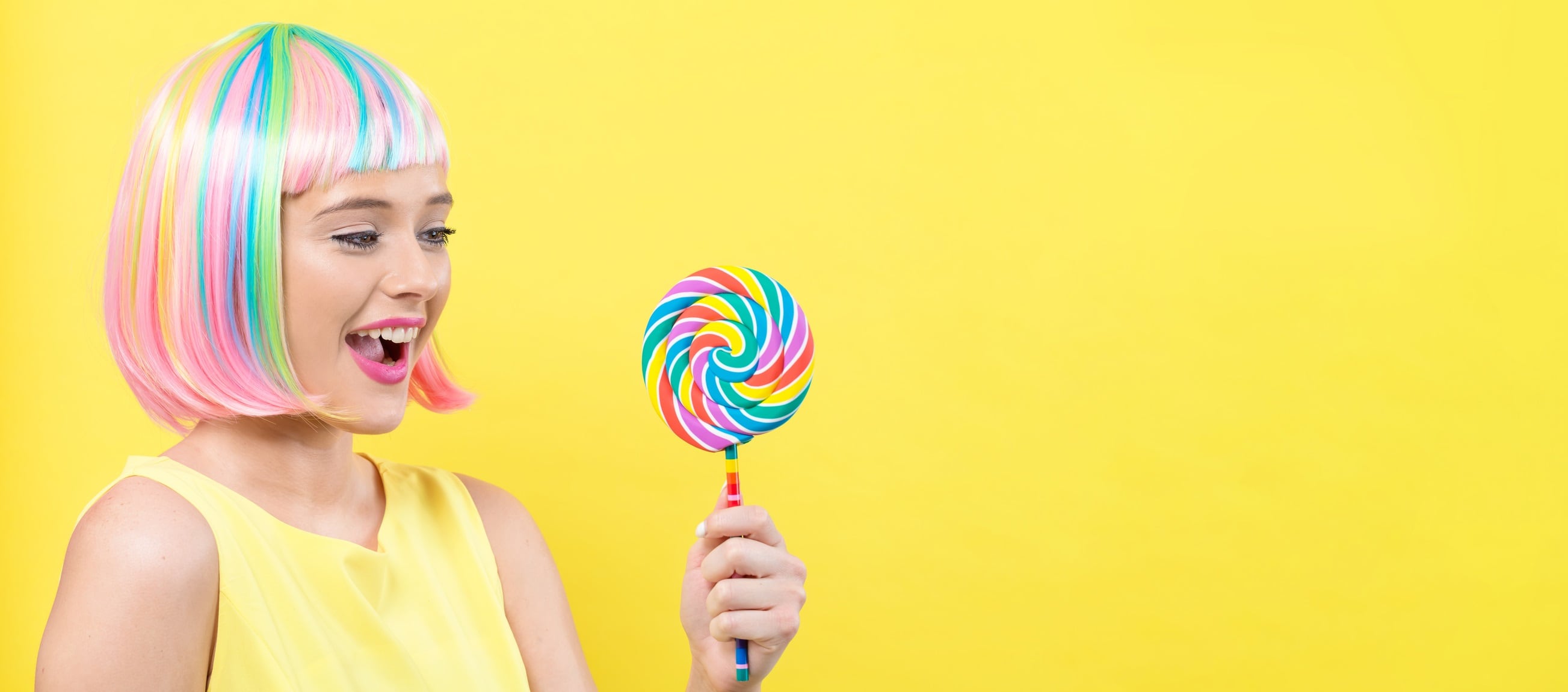 Woman with colourful hair, holding giant lollipop on a yellow background.
