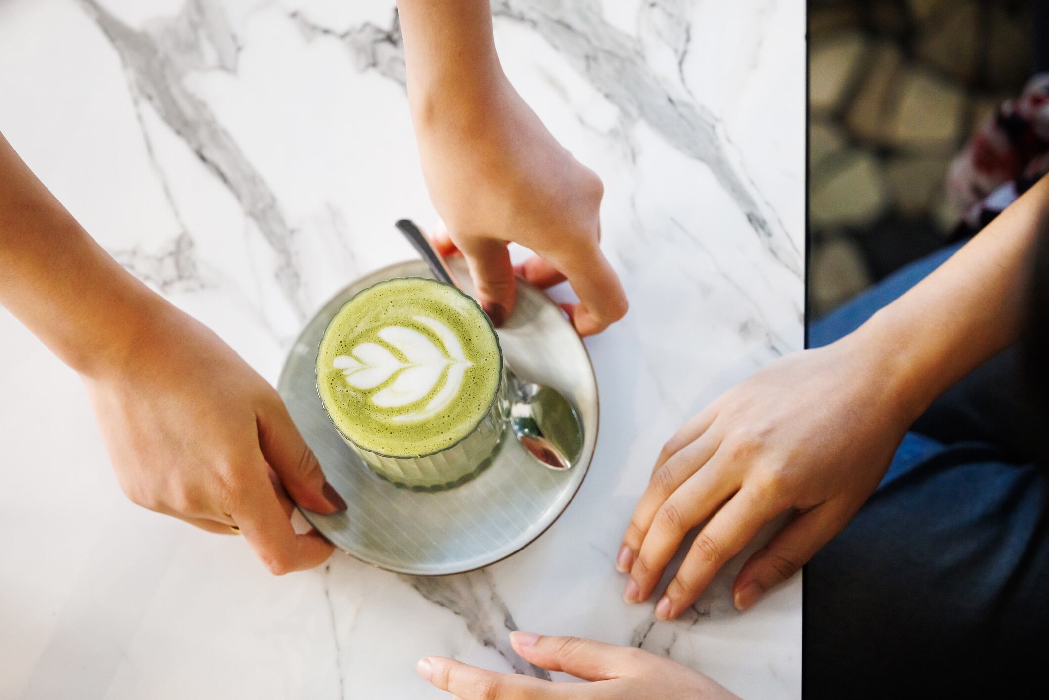 Overhead view of two hands reaching for a cup of matcha latte with artistic foam, placed on a marble surface.