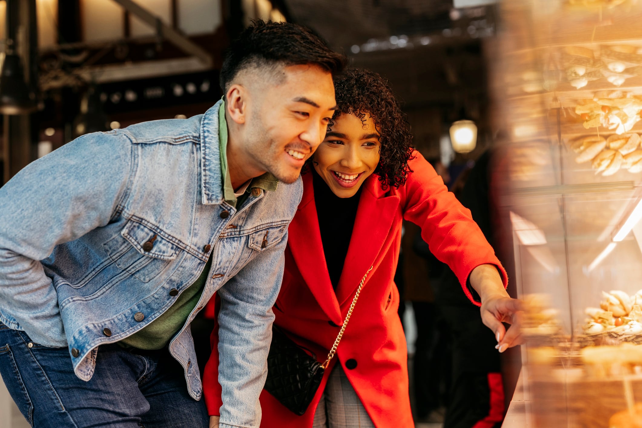 Young multiracial couple smiling and pointing while selecting delicious pastries in a bakery display window