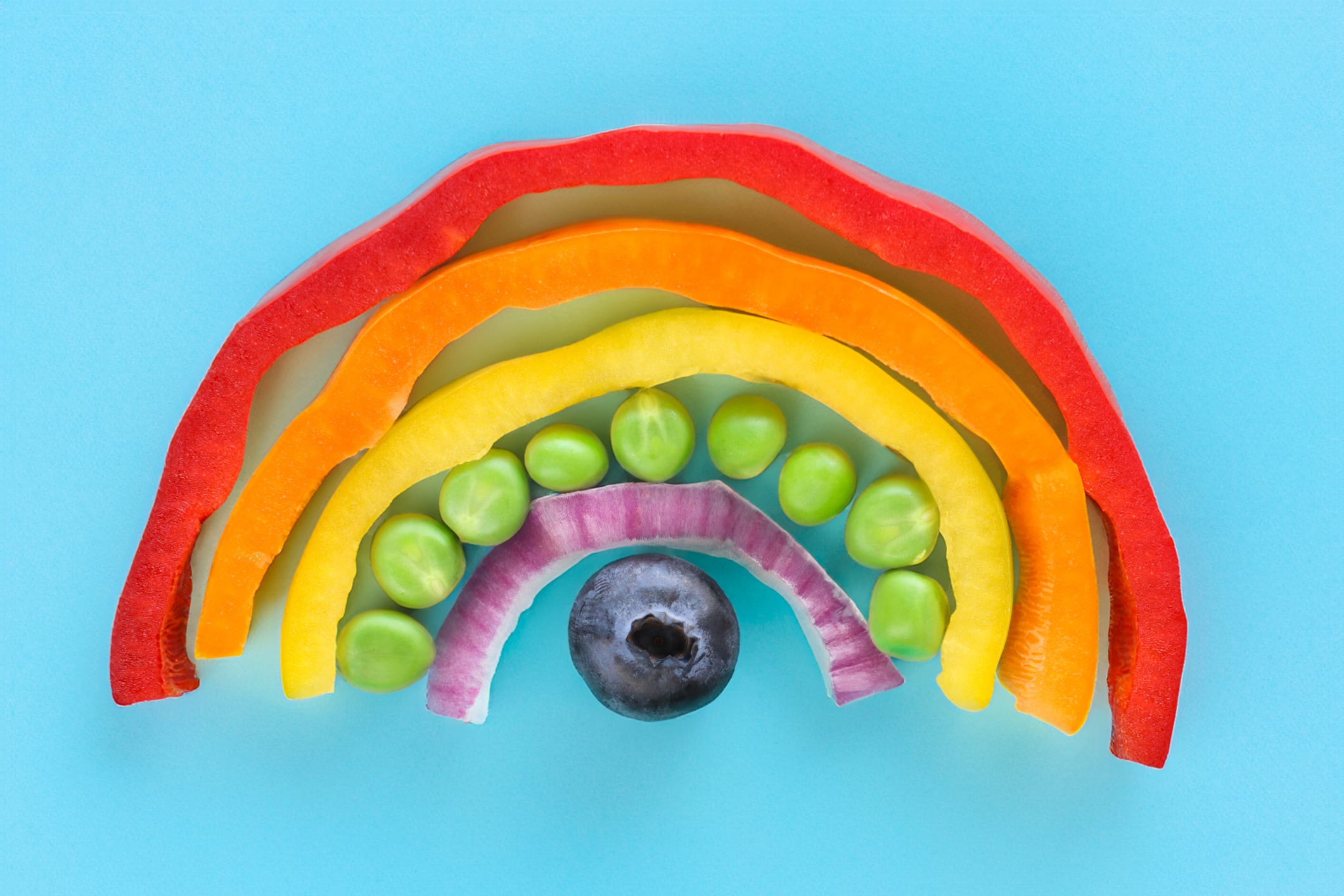Stock photo showing close-up, elevated view of arch of chopped multi-coloured fruits and vegetables. Rainbow of chopped red, orange and yellow bell peppers, green peas, red onion and a blueberry. Healthy eating concept.
