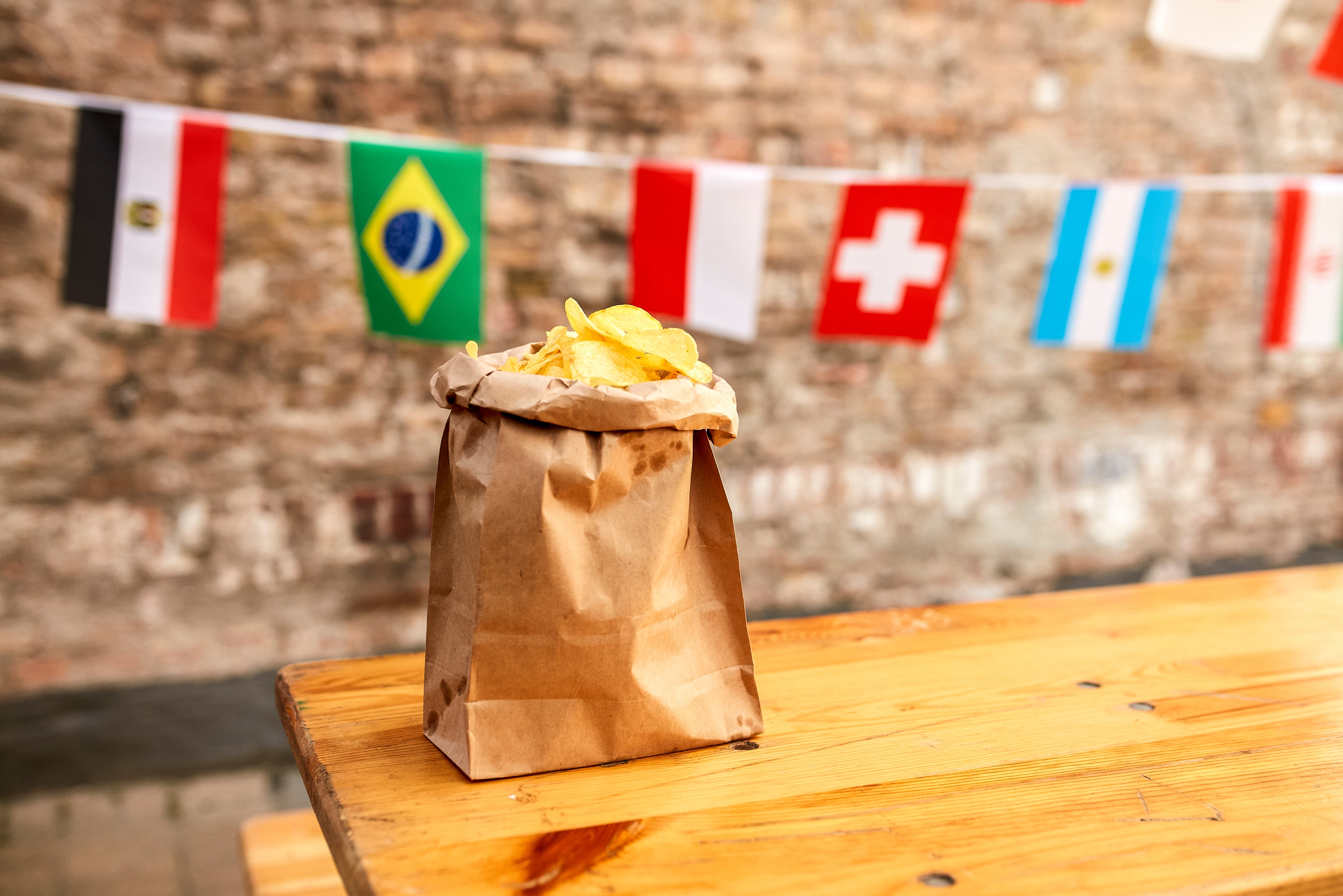A paper bag filled with potato chips is placed on a wooden table with international flags hanging in the background