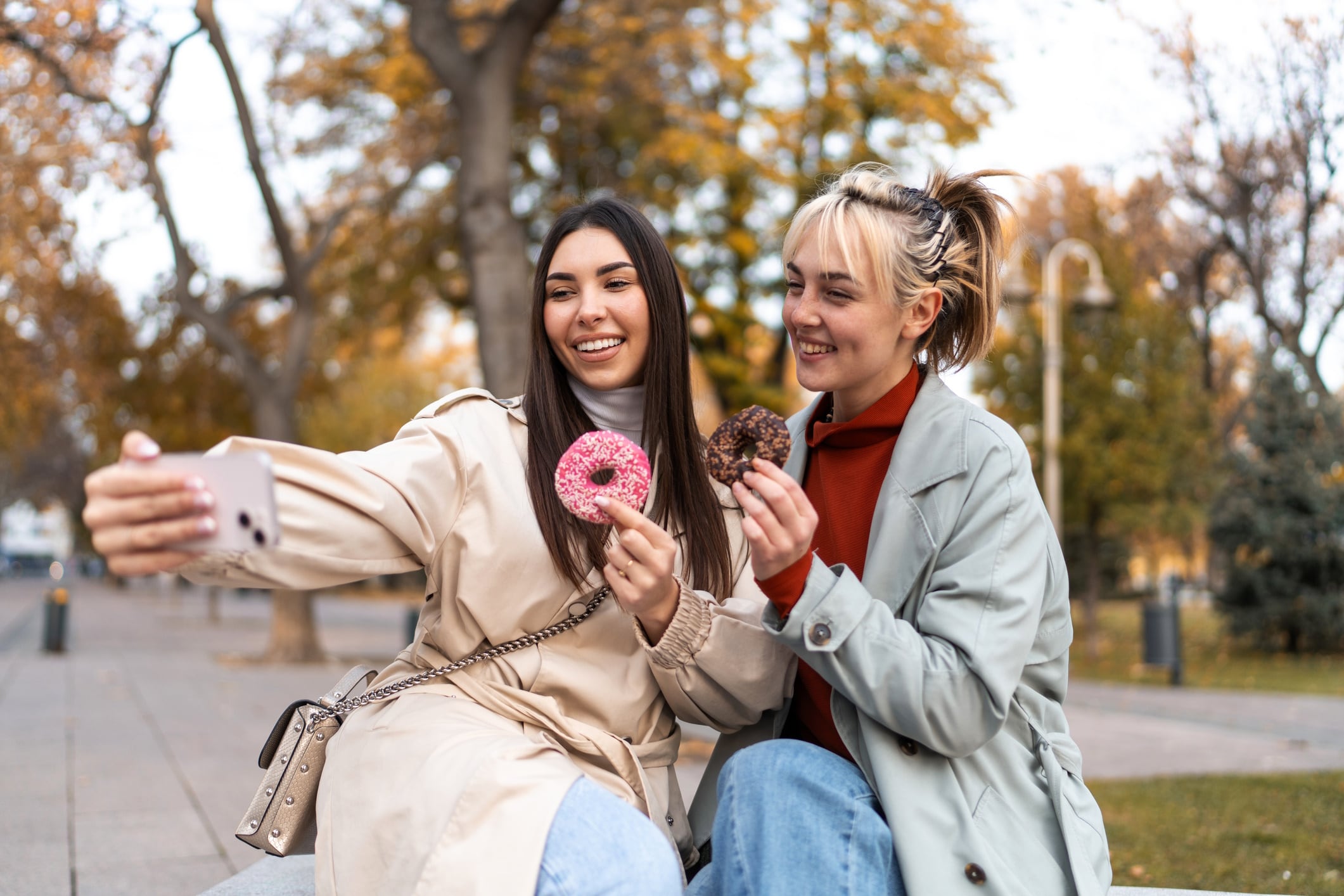 Two young women taking a selfie while eating donuts in a park