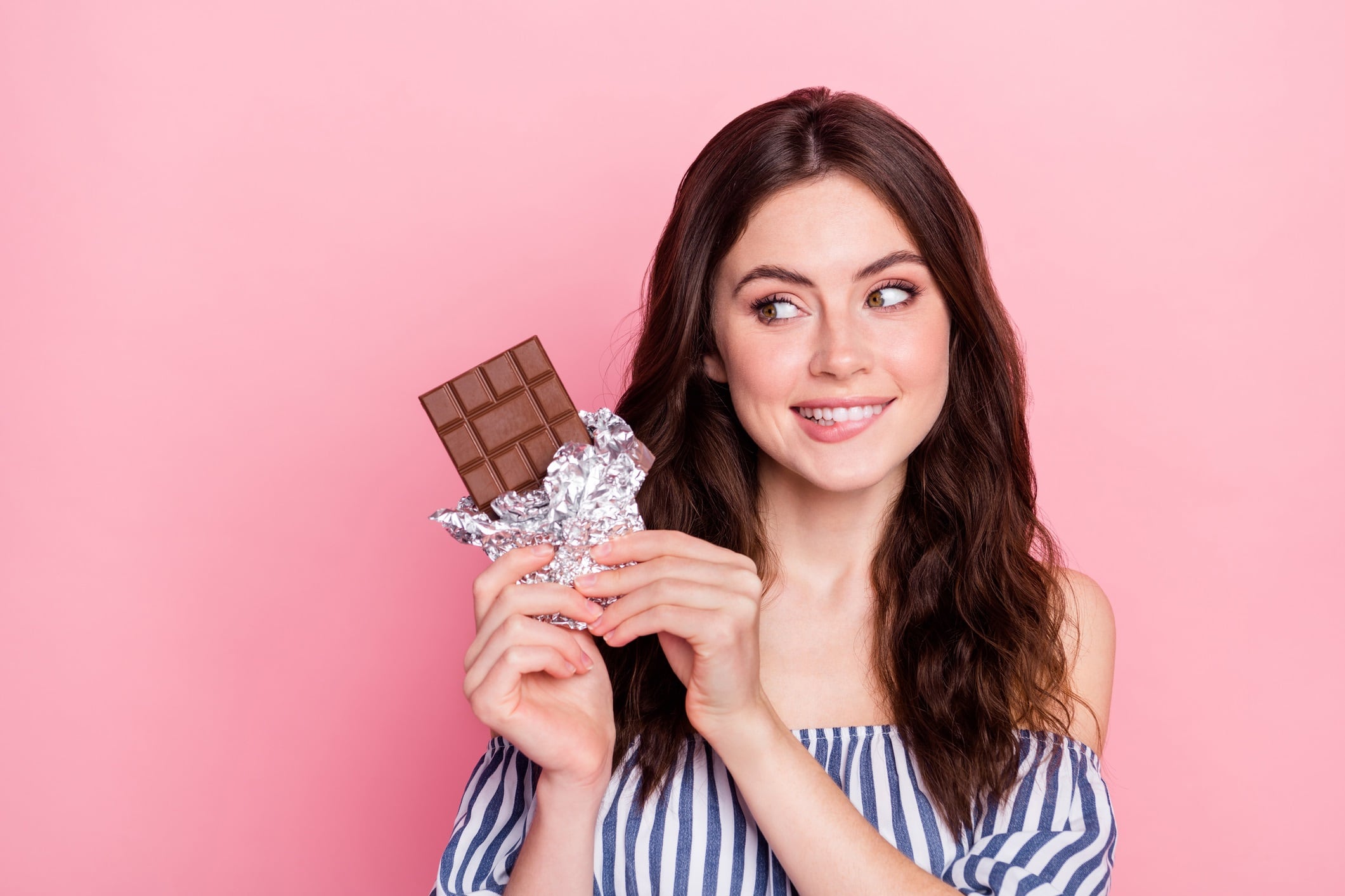 Woman wearing blue and white stripped shirt, holding chocolate bar. Pink-coloured background.