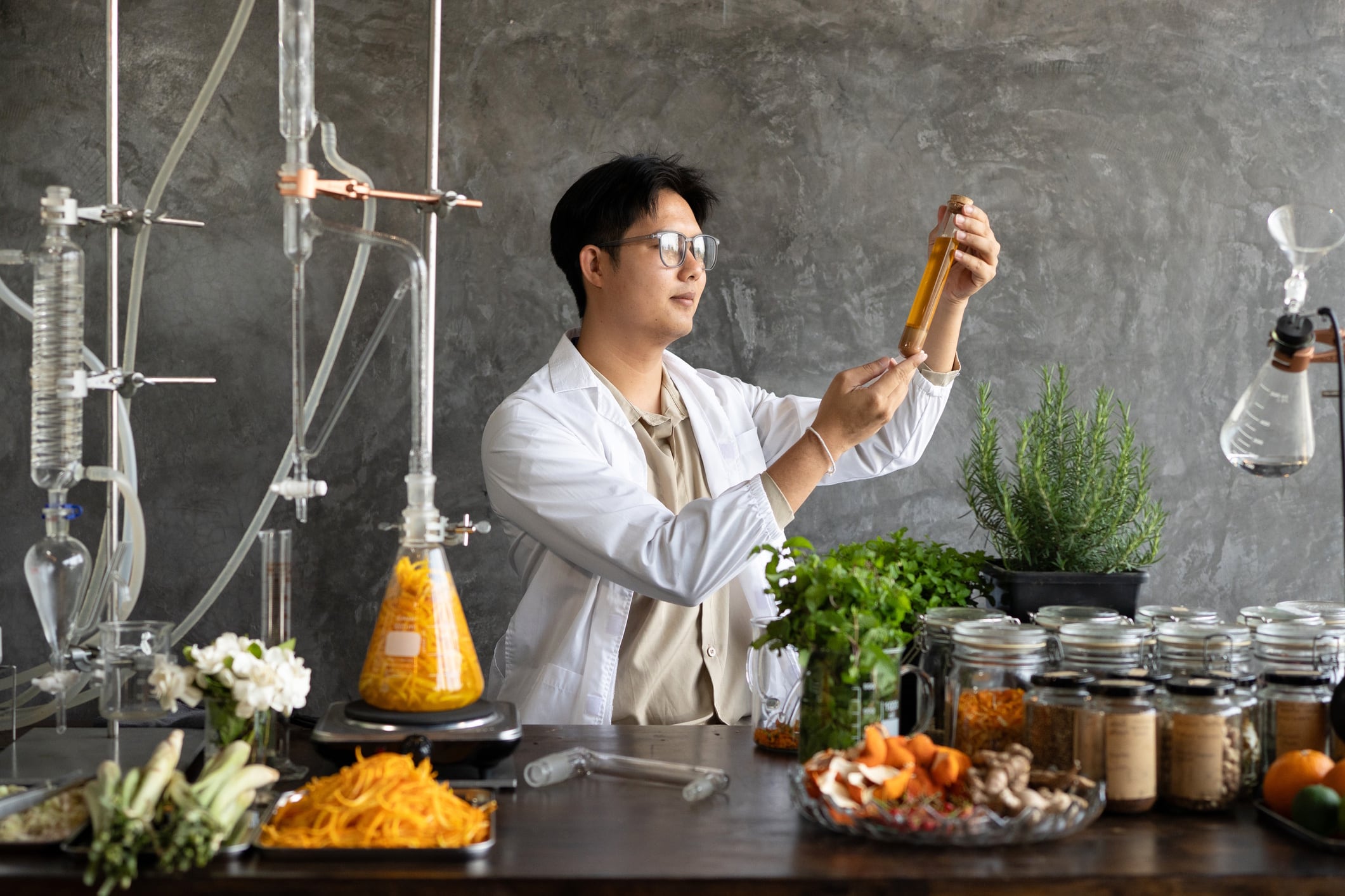 A focused Asian Scientist male researcher in a white lab coat inspects an amber liquid sample surrounded by herbs, spices, and glassware, ideal for natural medicine and wellness concepts Thidarat Kwangten GettyImages-