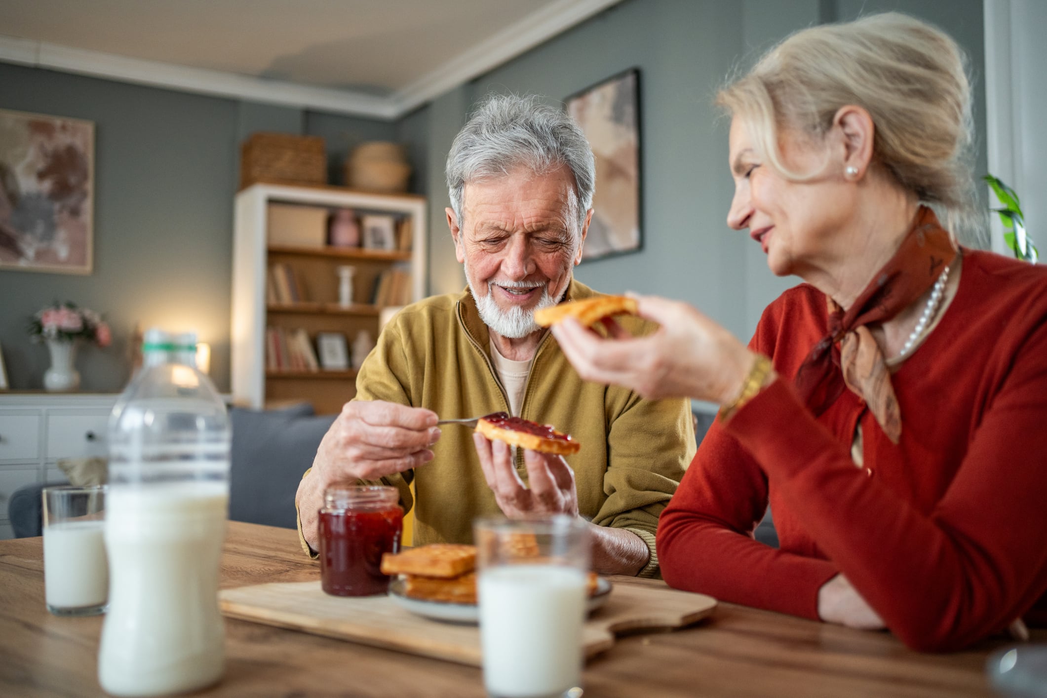 Elderly couple smiling and spreading jam on toast while having breakfast together at their dining table, enjoying a healthy and happy retirement