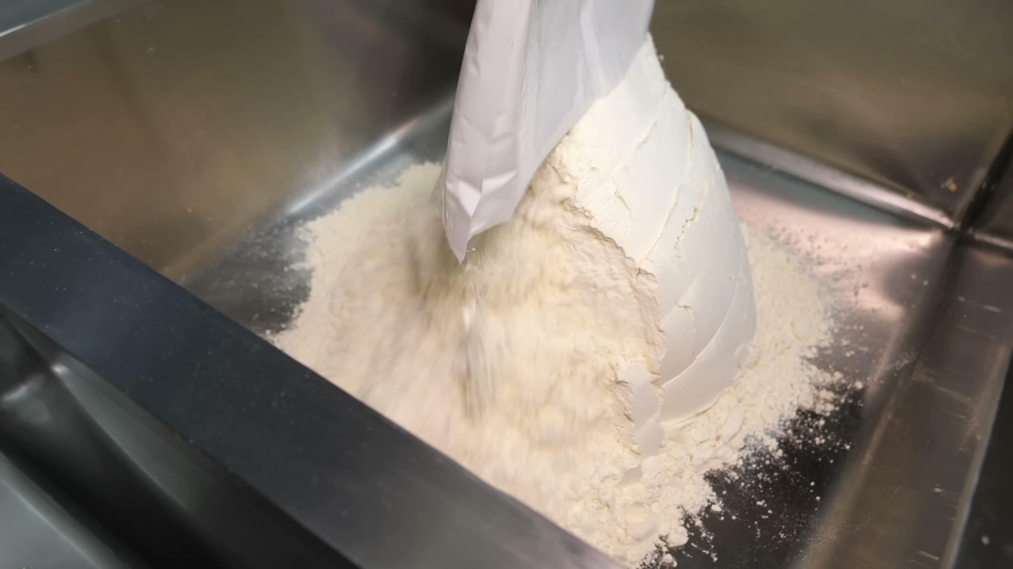 A person wearing black gloves pours a large quantity of white powdery flour from a plastic bag into a deep stainless steel industrial sink in a commercial kitchen while preparing bulk ingredients