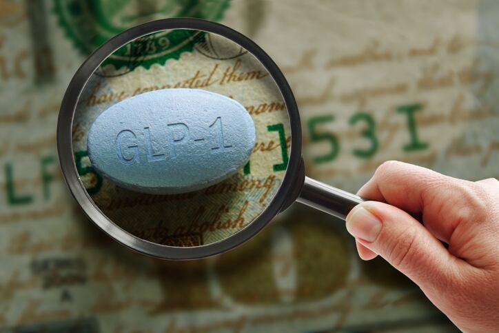 Hand holding a magnifying glass over a GLP-1 pill on a hundred dollar bill