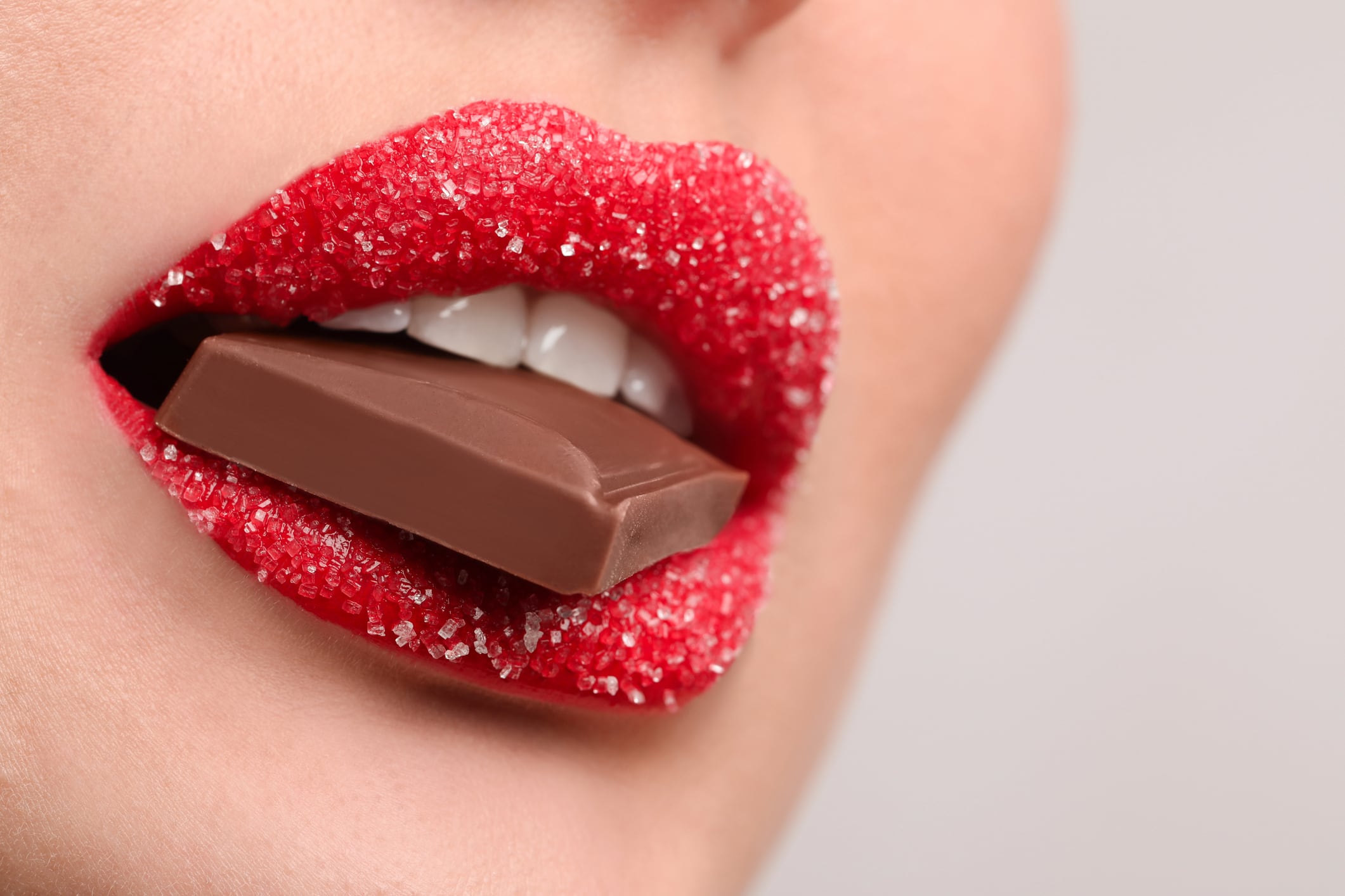 Woman wearing red lipstick, with sugar on, biting chocolate. Light grey background.