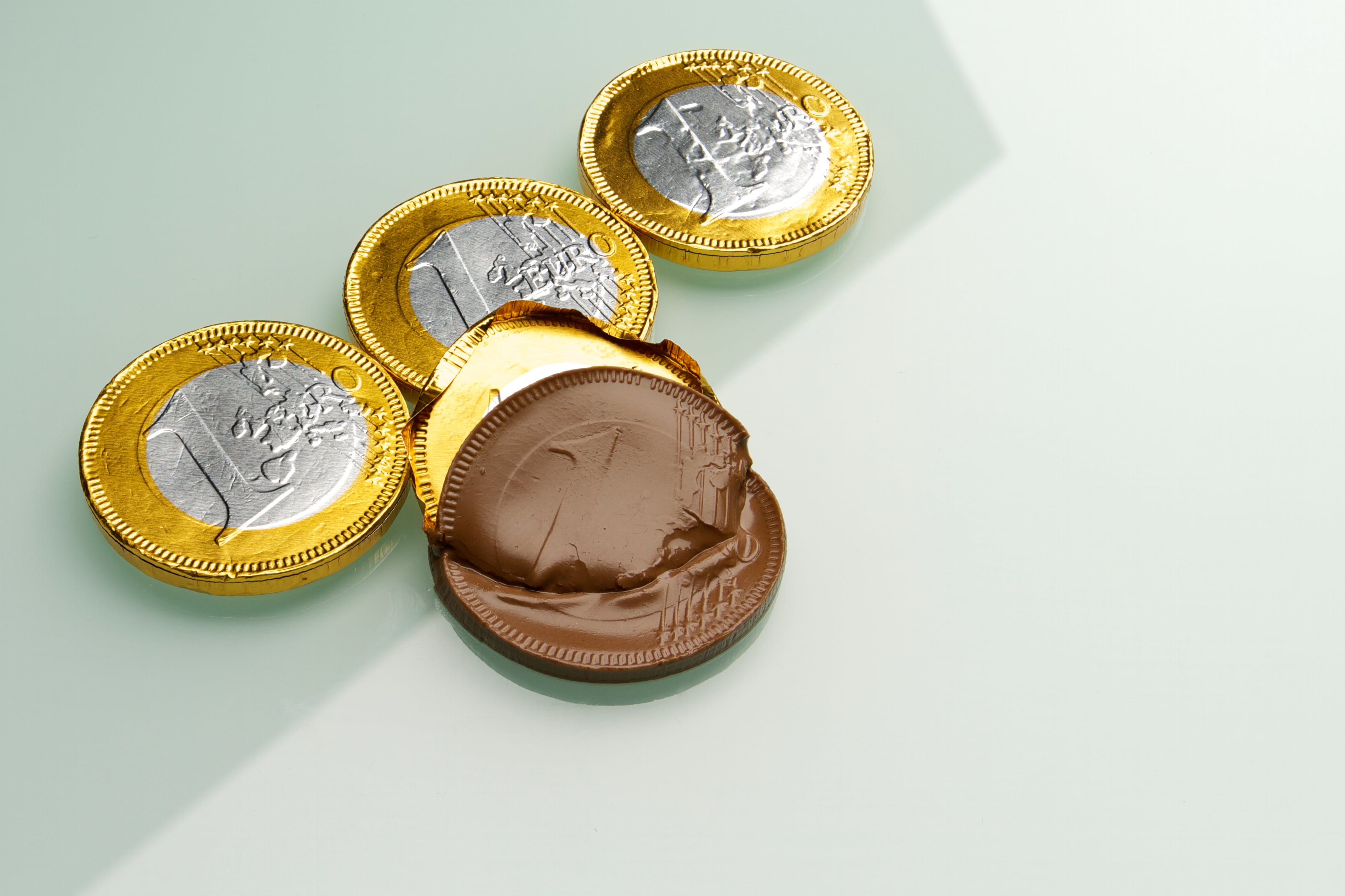 Four chocolate Euros, one opened and melting, set on a reflective, white base