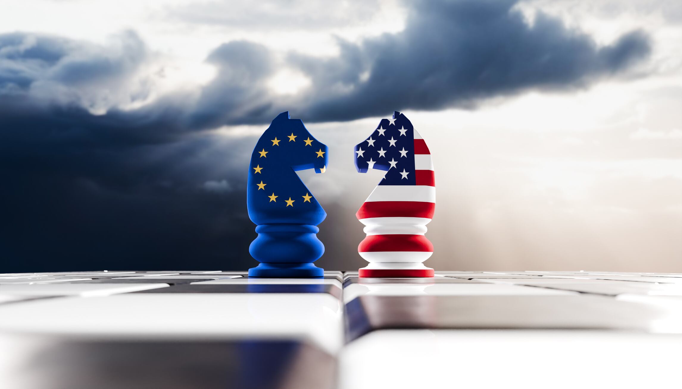 Two rooks stand poised in a face-off, each showcasing the colors of the American and European Union flags, set against a moody, overcast sky. The striking reds, whites, and blues of the U.S. flag contrast with the deep blue and gold of the EU flag, symbolizing the intricate relationship and rivalry between the two powers. The dark, tumultuous clouds above amplify the sense of tension and uncertainty, evoking themes of diplomacy, competition, and the strategic maneuvering often seen in international relations.