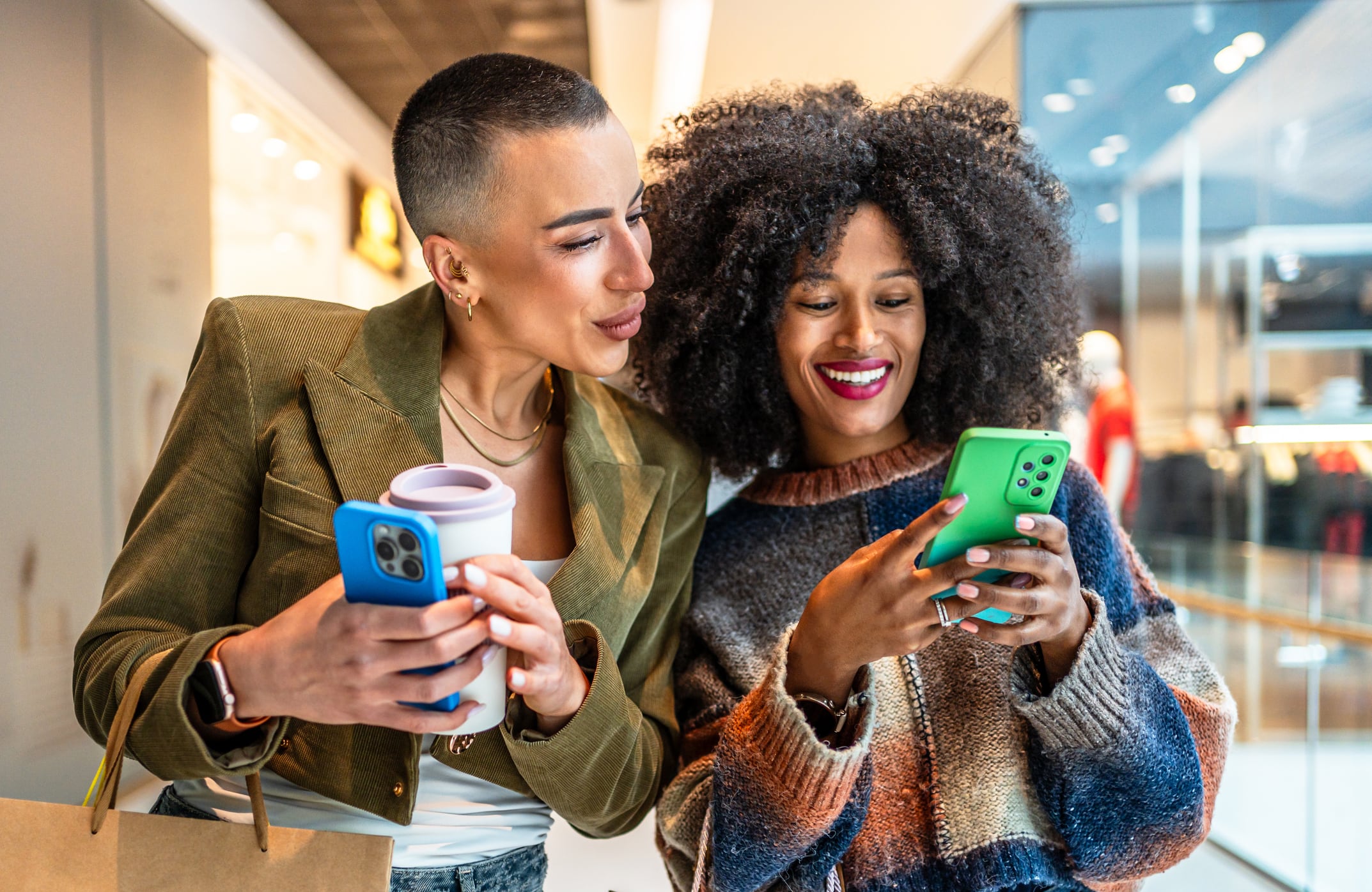 Two joyful female friends enjoying shopping in a mall, sharing moments while browsing on a mobile phone and sipping coffee together