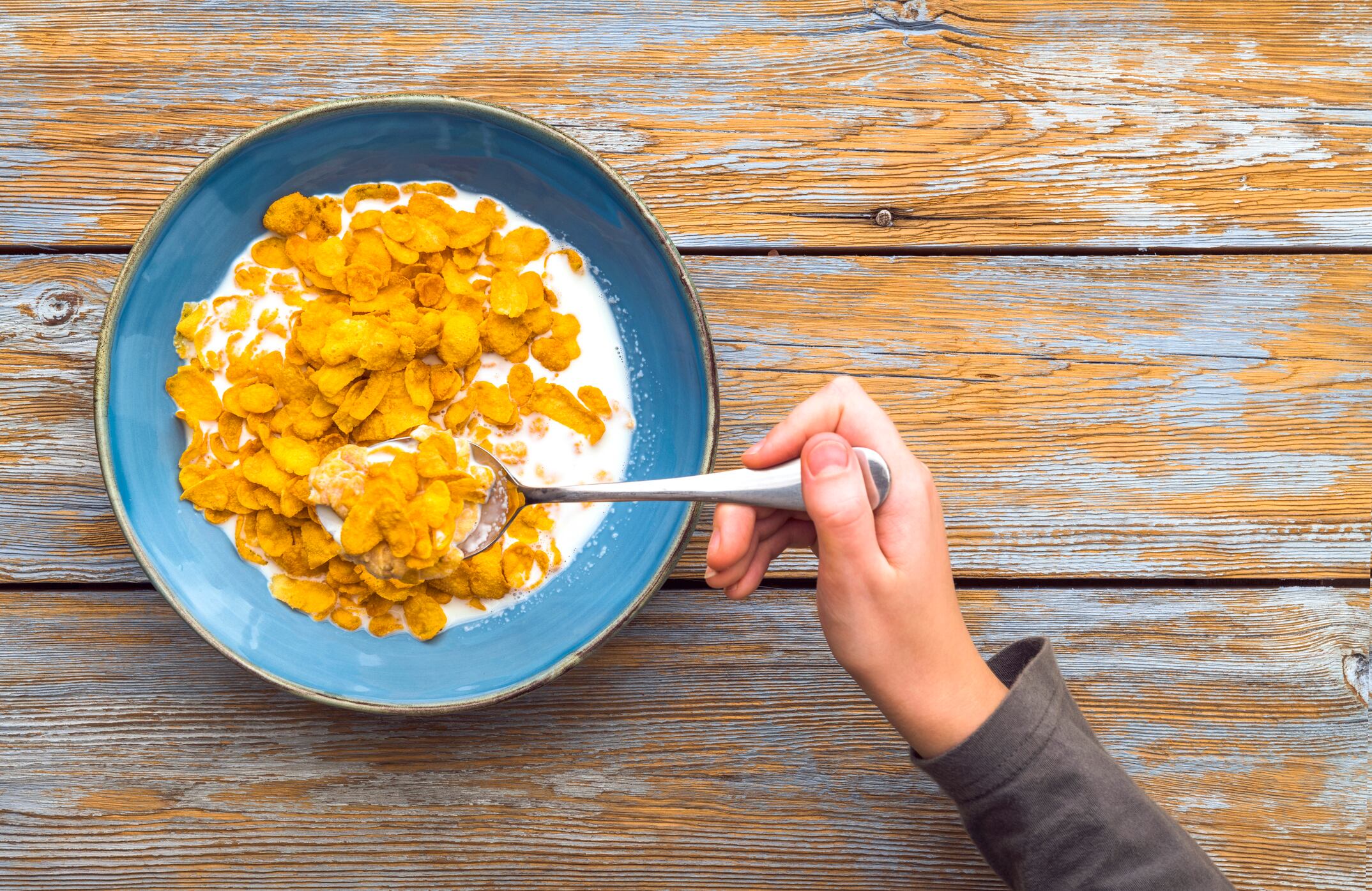 Child eating corn flakes for breakfast on wooden table. Top view