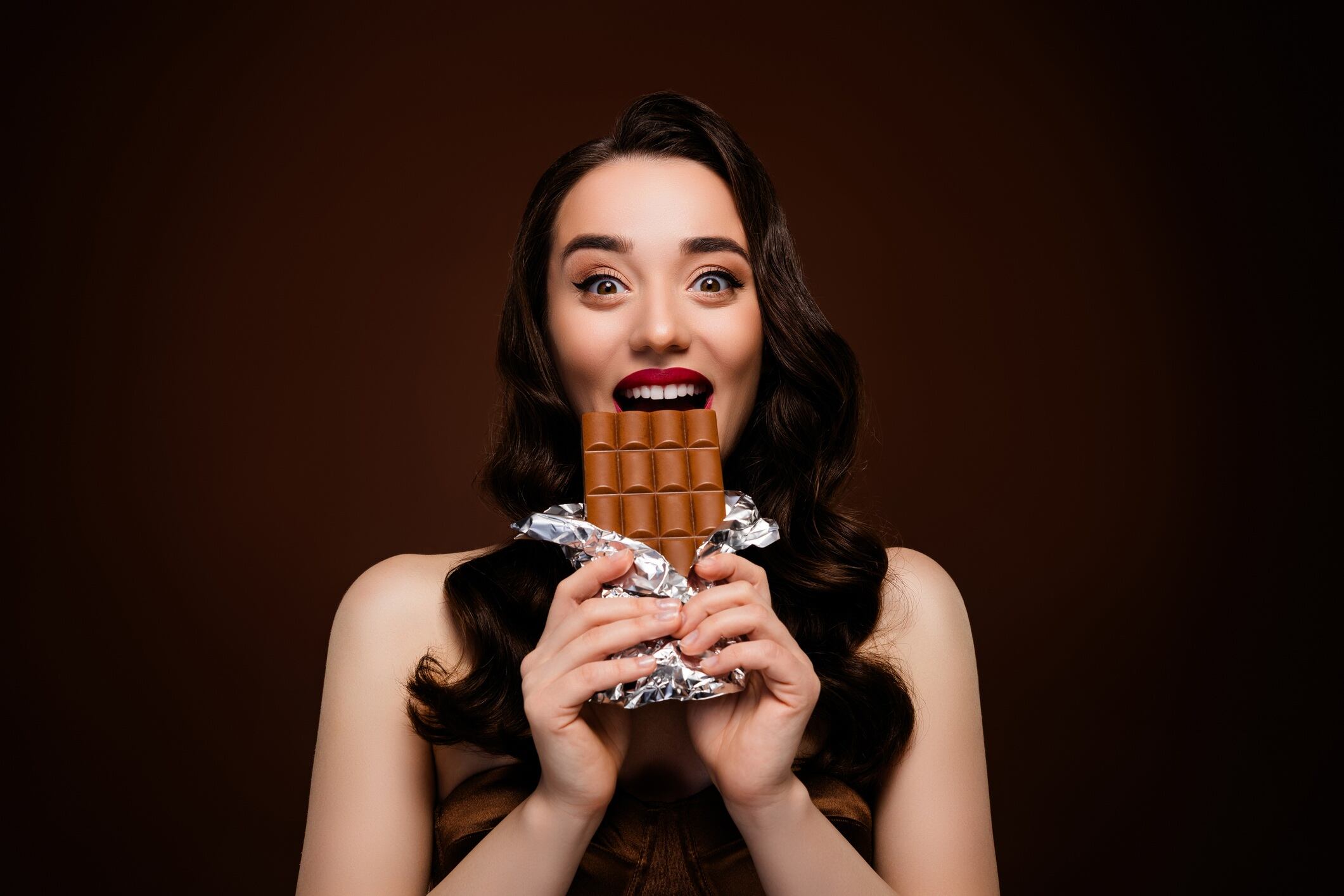 Woman wearing red lipstick and smiling, holding chocolate bar. Isolated on brown coloured background.
