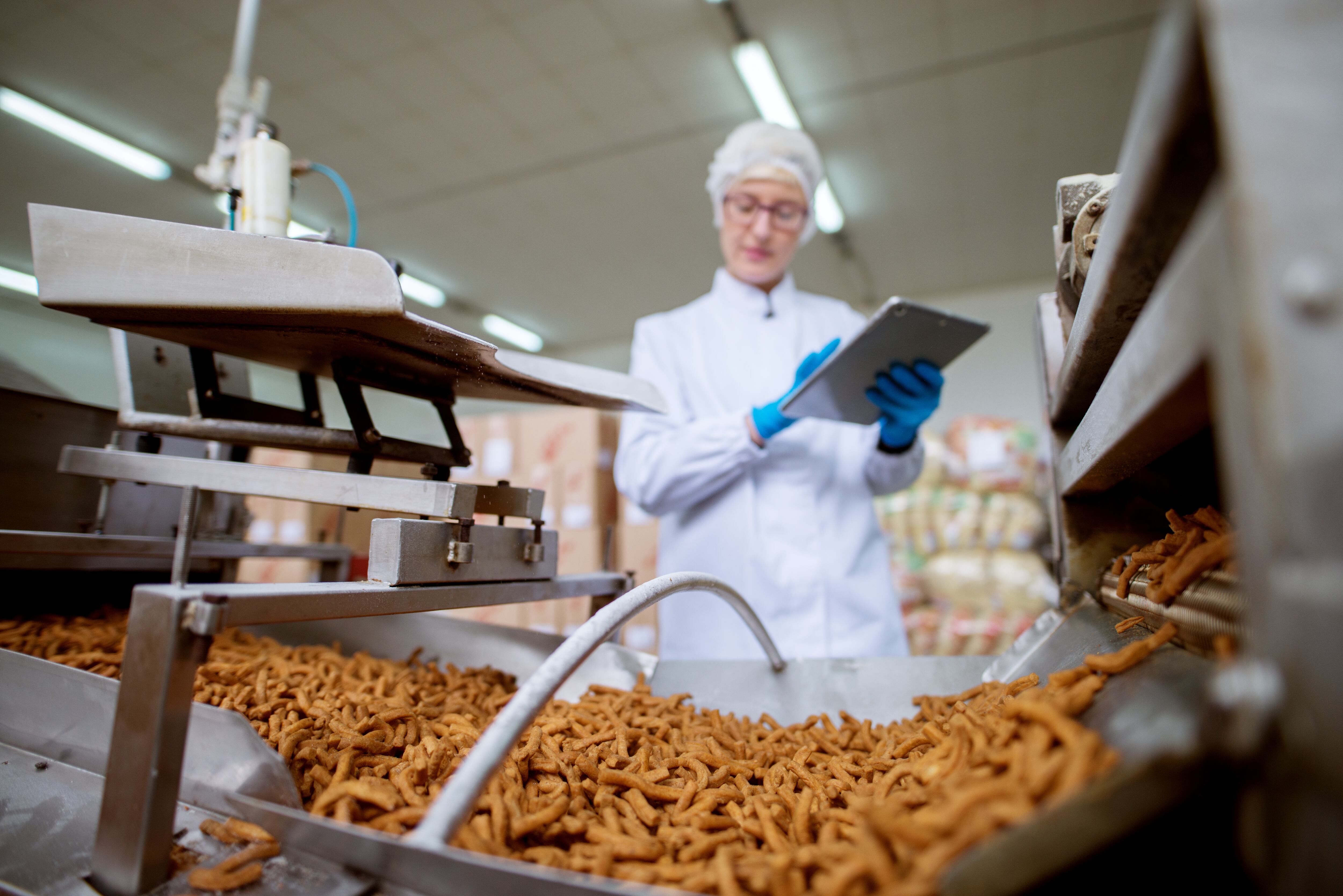 Close up view of a salt stick snacks being processed through a production line in food factory while being inspected by a female young worker with a tablet in a sterile cloth. dusanpetkovic GettyImages