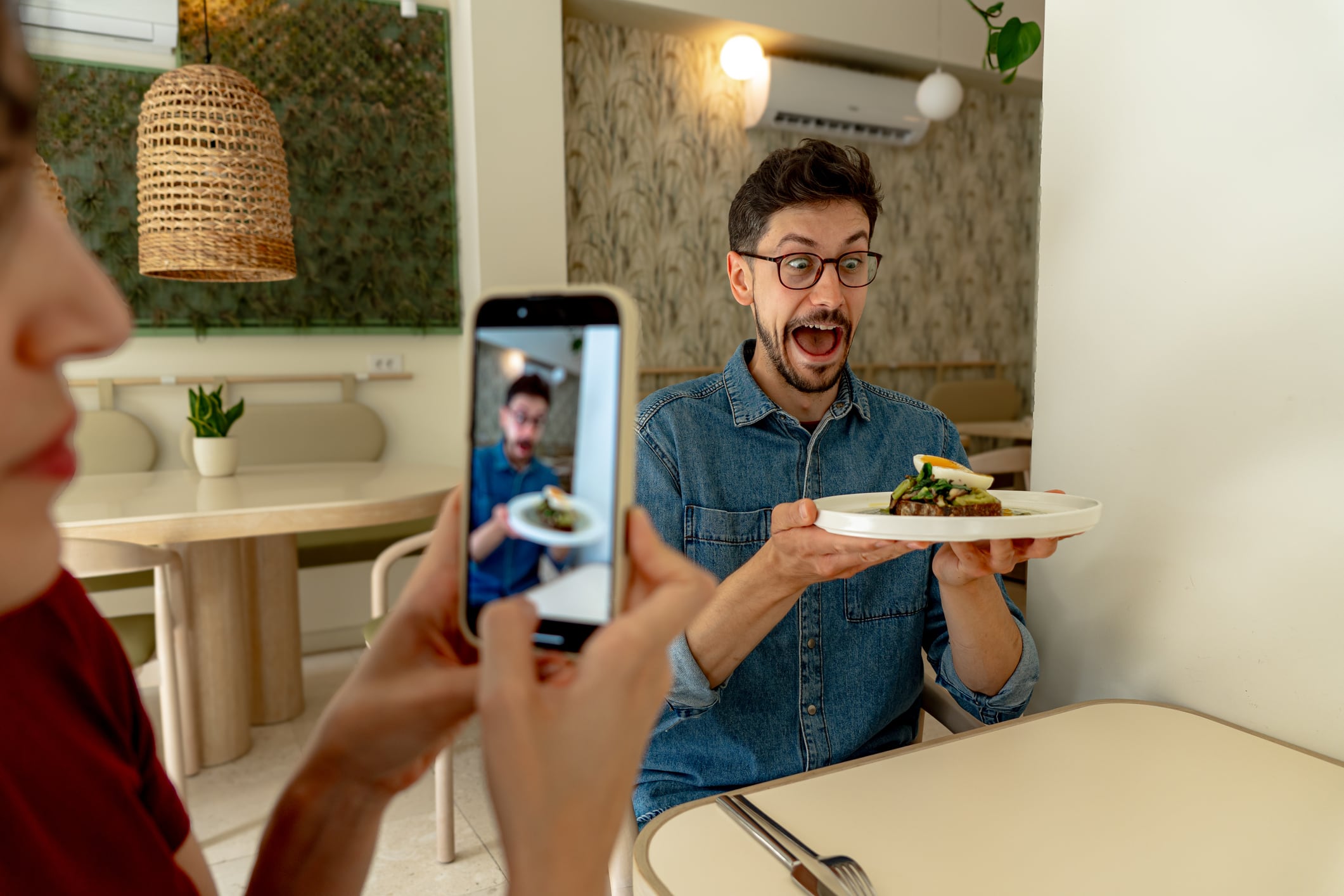 Man showing his gourmet dish with surprised expression while woman taking photo with phone in modern restaurant, sharing food experience