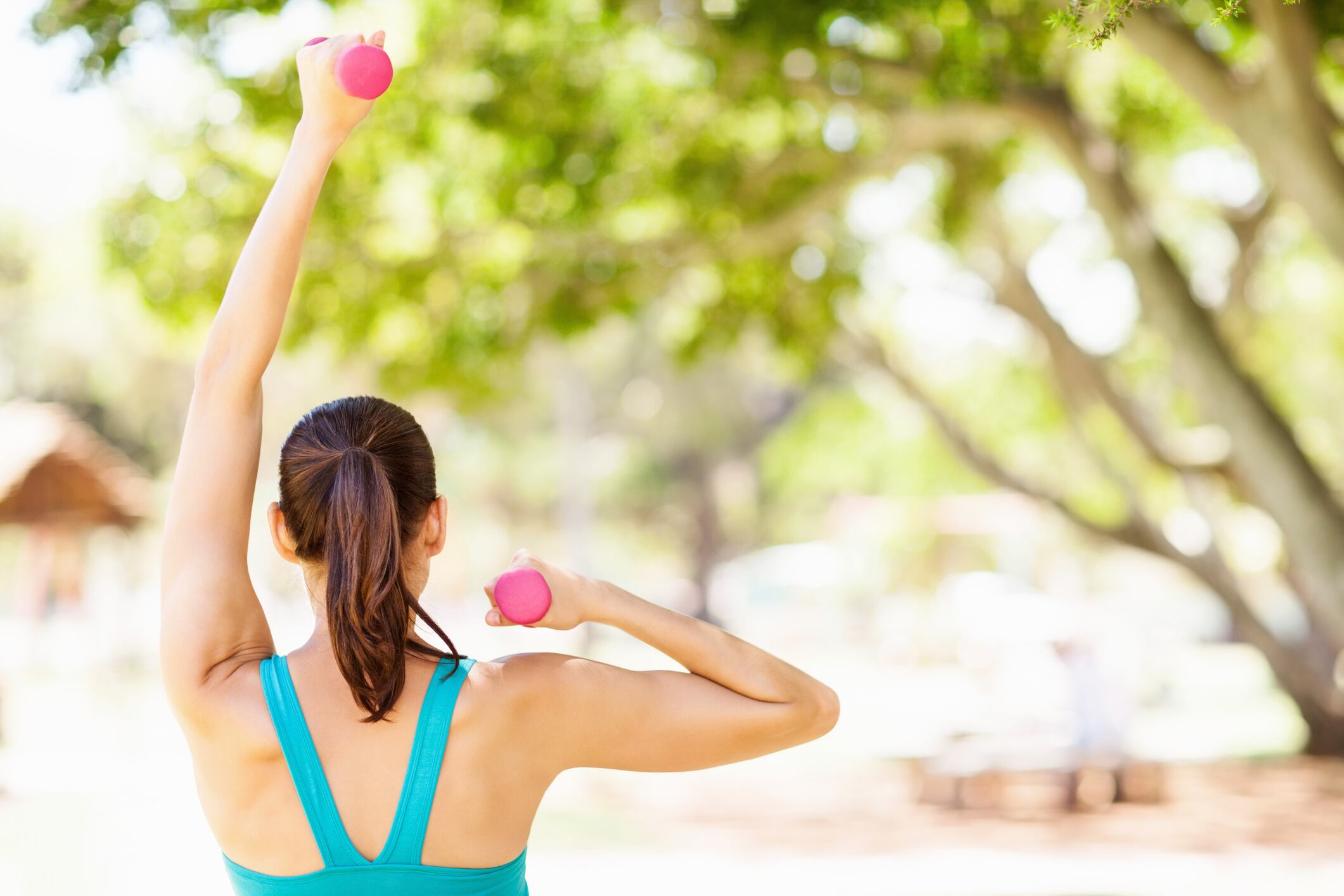 Rear view of sporty young woman lifting dumbbells in park. Horizontal shot.