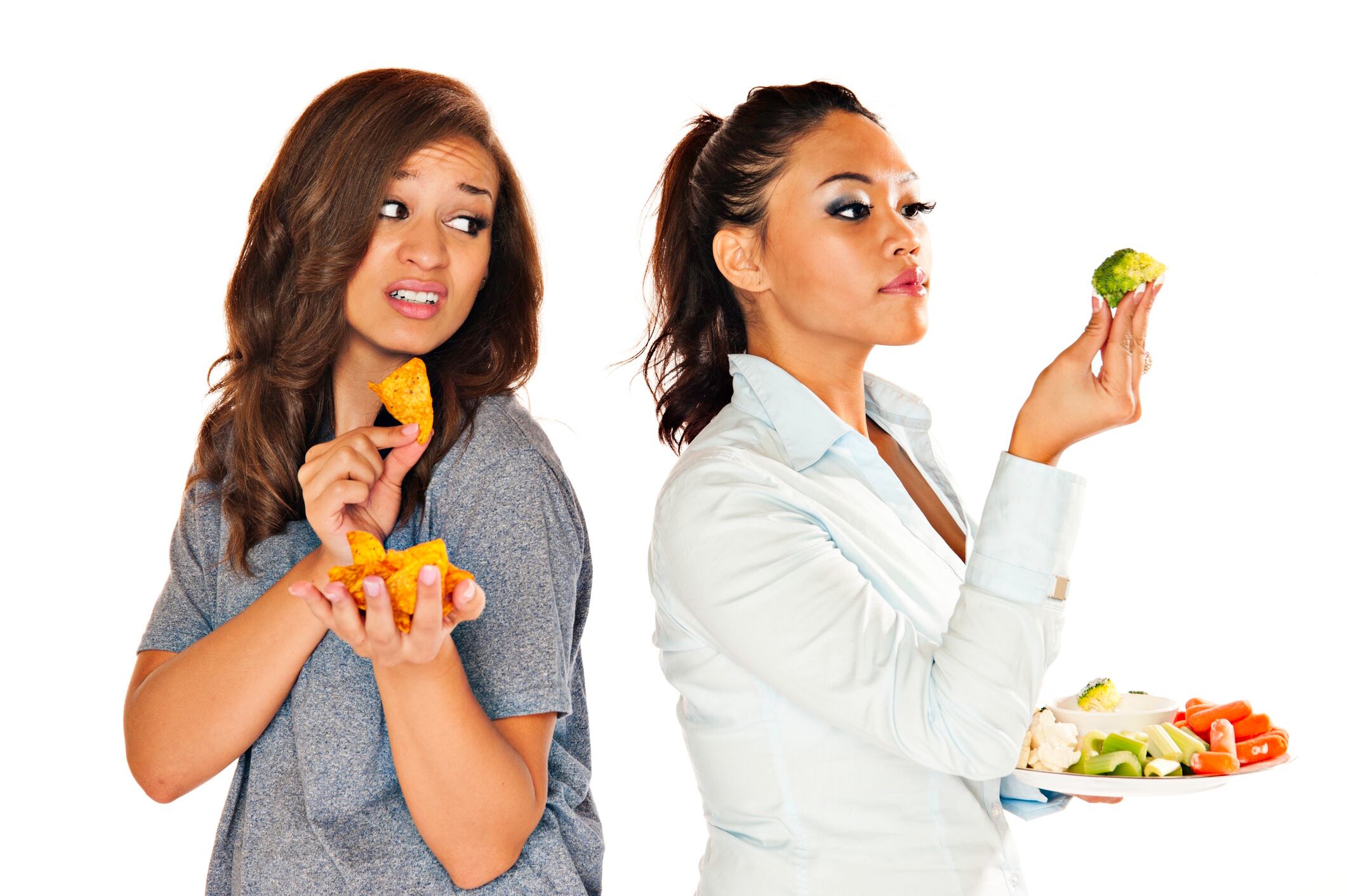 Two women with one holding up healthy vegetables and the other holding chips demonstrating the daily challenge of eating healthy. InkkStudios GettyImages