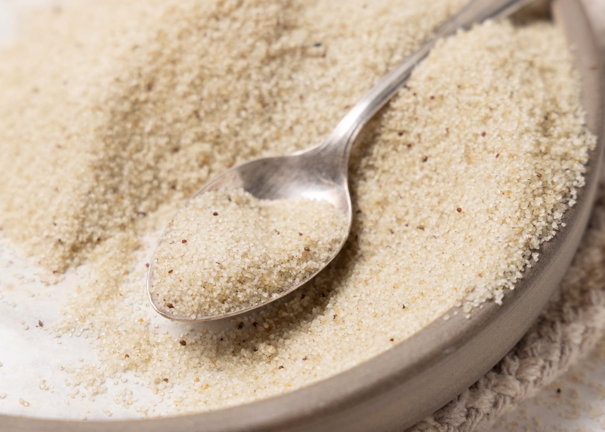 Raw uncooked fonio seeds in a plate with a metal spoon on white table close up. African ancient pseudocereal. Organic super food. Healthy food concept. Gluten free alternative seeds