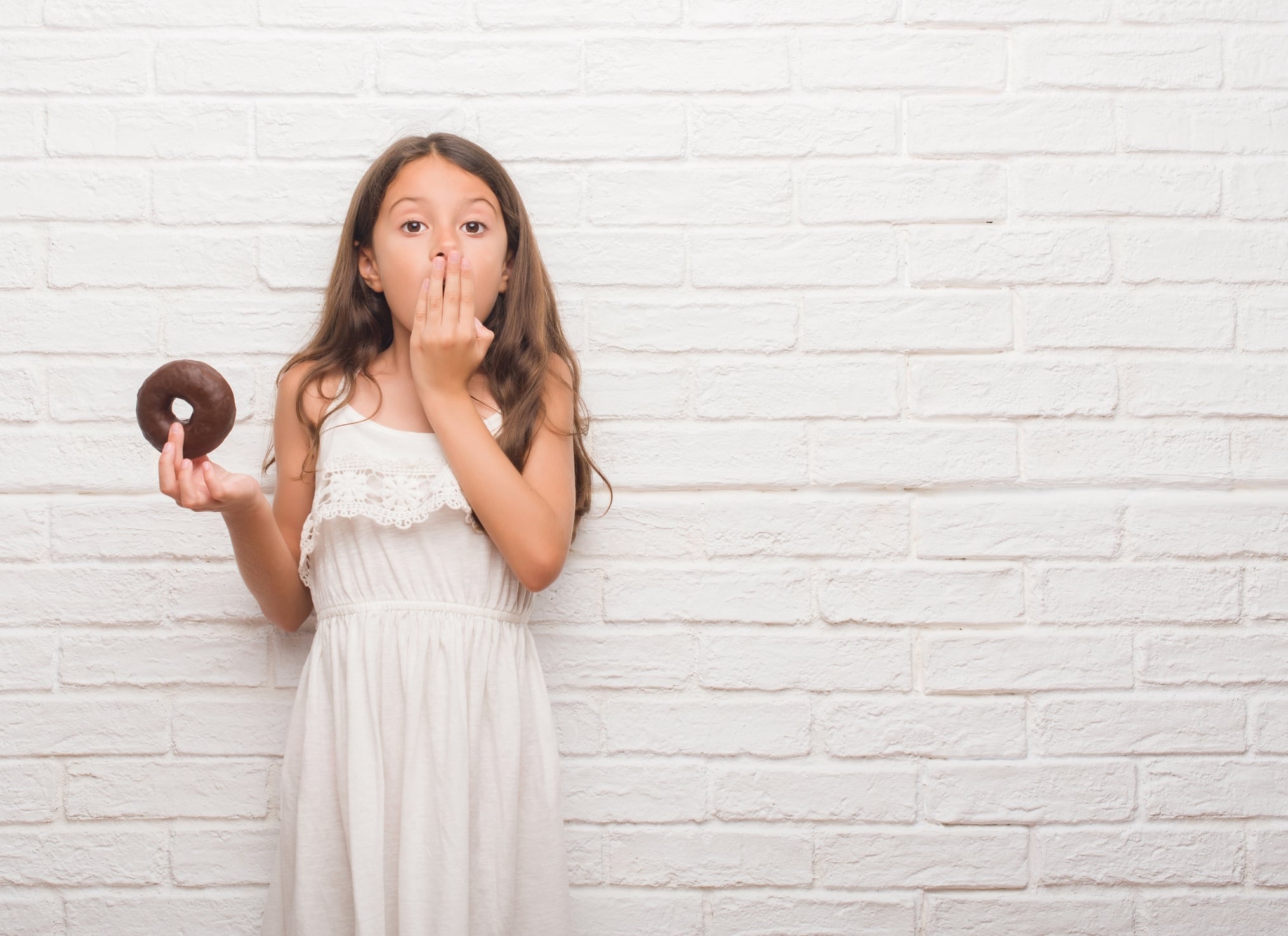 Young hispanic kid over white brick wall eating chocolate donut cover mouth with hand shocked with shame for mistake, expression of fear, scared in silence, secret concept