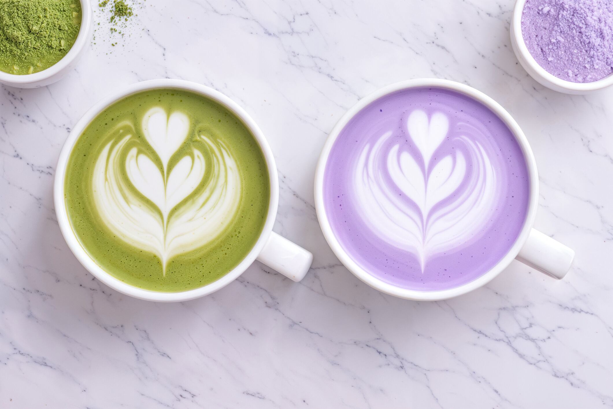 Macro top view of two colorful café beverages placed side by side: a vibrant ube latte and a green matcha latte, both topped with delicate latte art. This visually striking comparison highlights the contrast between the soft purple tones of ube purple yam and the rich green color of matcha tea. Both ingredients are widely appreciated in modern cafés for their natural origin, antioxidant properties, and association with wellness-focused lifestyles. The smooth foam textures and symmetrical composition create an elegant presentation ideal for contemporary coffee culture. This aesthetic beverage scene is perfect for café menus, specialty drink promotions, food photography, and social media content.