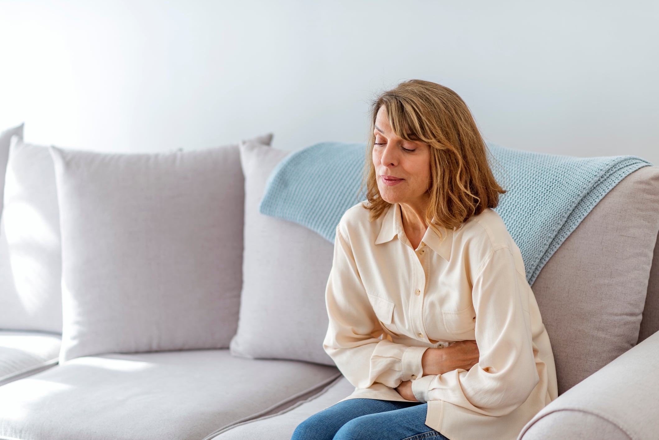 Senior woman suffering from stomach ache sitting on a bed in the bedroom. Photo of Senior woman sitting on her bed has a stomach ache in the bedroom. Senior woman suffering from stomach pain while sitting on a gray sofa during the day.