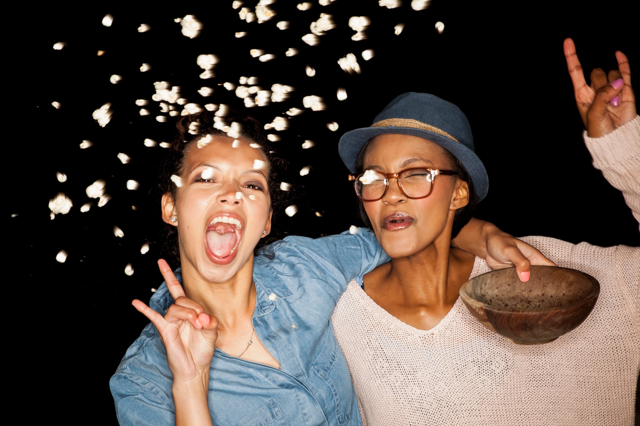 Young women throwing popcorn from bowl looking at camera doing rock sign DUEL GettyImages