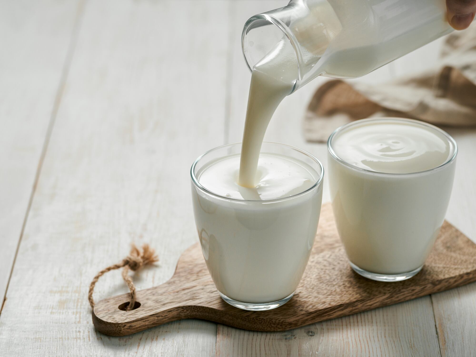 Pouring homemade kefir, buttermilk or yogurt with probiotics. Yogurt flowing from glass bottle on white wooden background. Probiotic cold fermented dairy drink. Trendy food and drink. Copy space left