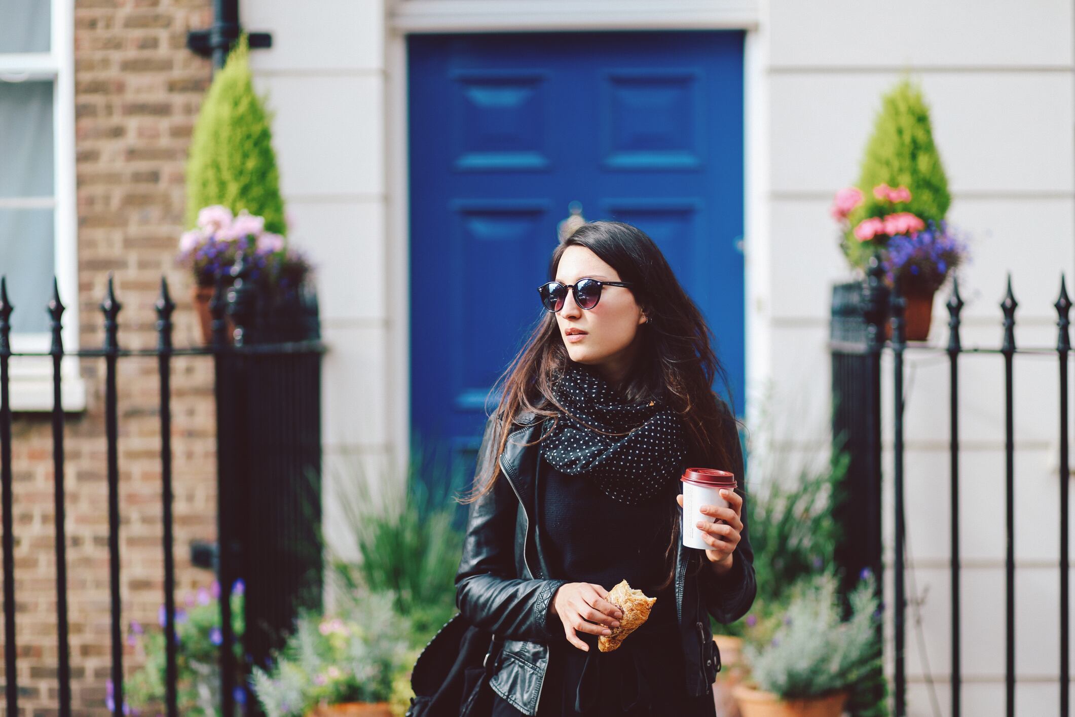 Woman, walking in London. She's wearing a black leather jacket and sunglasses, and holding a coffee and sandwich