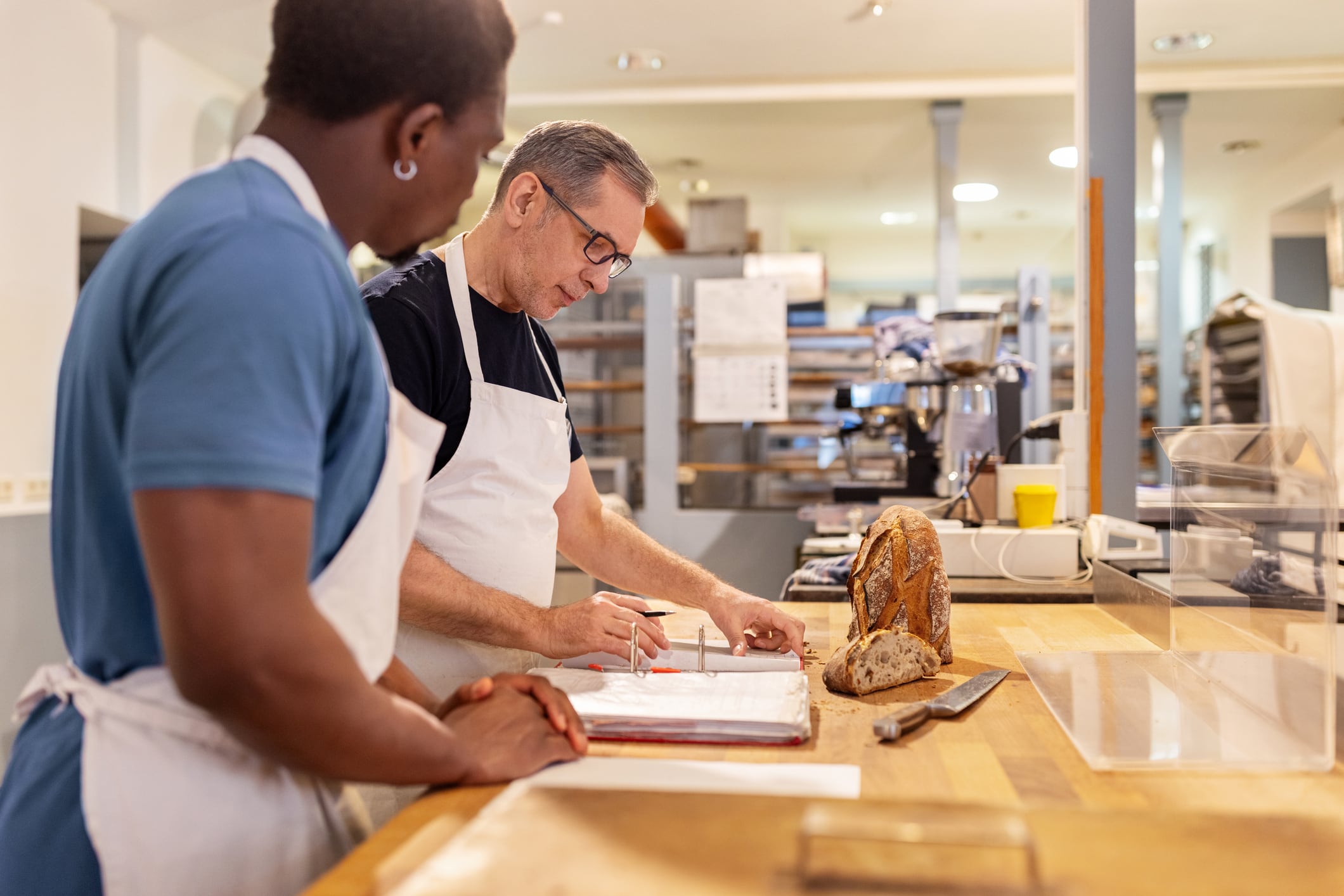 Senior baker mentoring new employee bakery finances. Experienced senior baker demonstrating to a new employee how to handle accounts and finances in a bakery. Mentorship, training, and the financial management aspects of running a bakery.