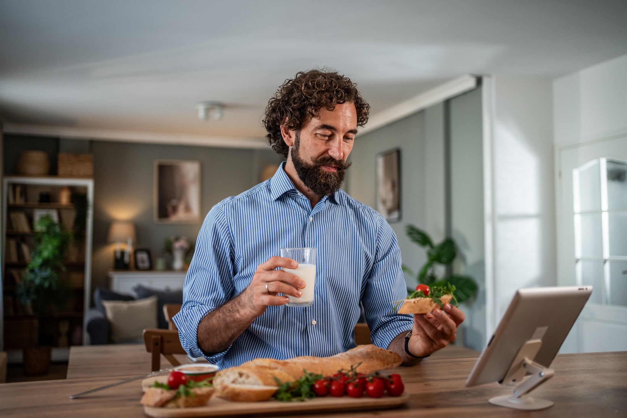 Mid adult man enjoying a nutritious breakfast of bread, tomatoes, and milk at home, looking at a digital tablet for entertainment or news during his morning routine